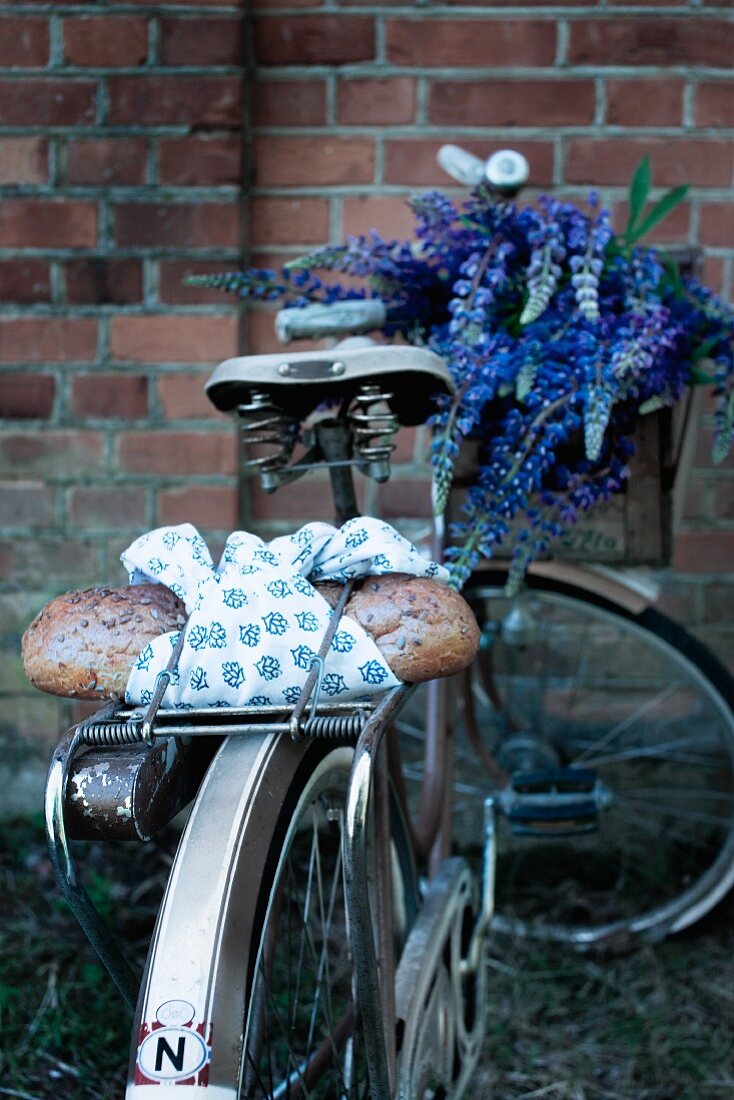 In blauweiss gemustertes Tuch gewickeltes Brot auf dem Gepäckträger eines Fahrrades mit Lupinenstrauss in Lenkerkiste vor Backsteinmauer