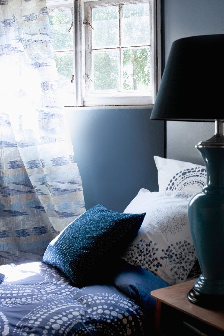 Blau gemusterte Kissen und Decke auf Bett vor Sprossenfenster mit Gardine