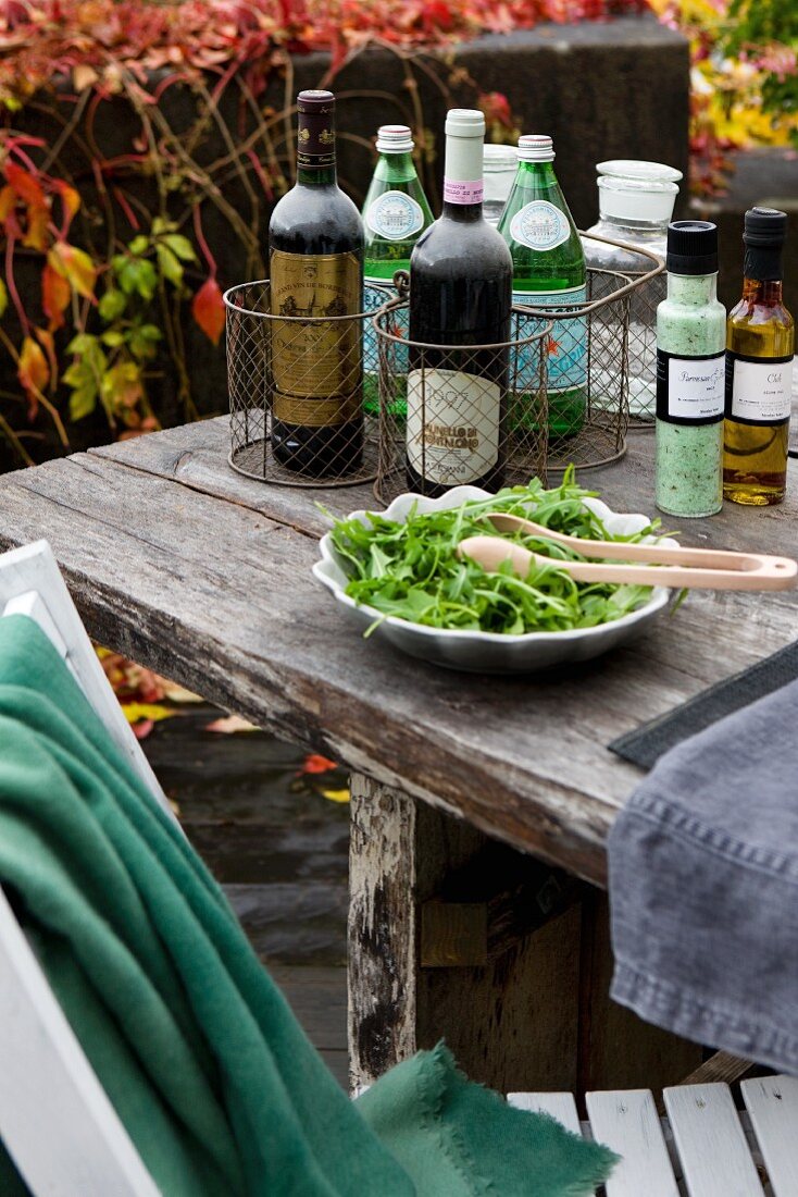 Weinflaschen in Drahtkörben, Salatteller und Dressing-Flaschen auf rustikalem Tisch; herbstliches Weinlaub im Hintergrund