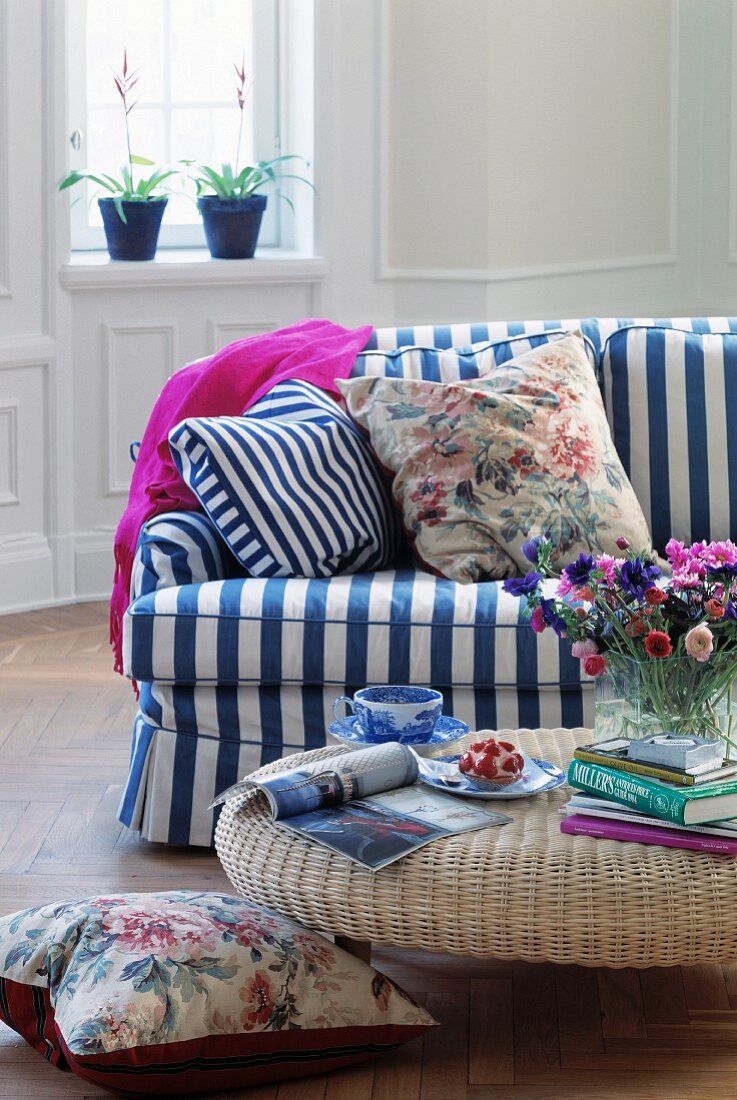Wohnzimmer mit weisser Wandvertäfelung und einer weiss-blau gestreiften Couch; im Vordergrund ein runder Rattan-Couchtisch