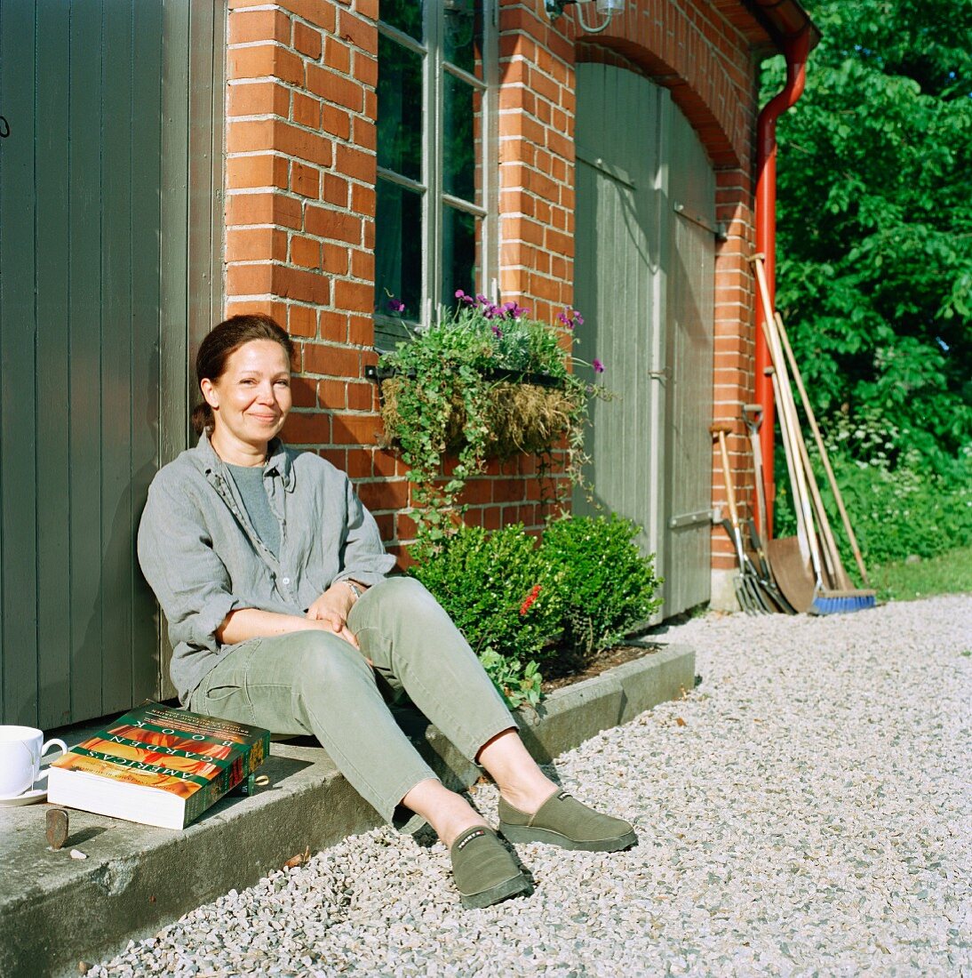 Frau sitzt mit Buch & Kaffeetasse in der Sonne auf Stufe vor Tür im Hof