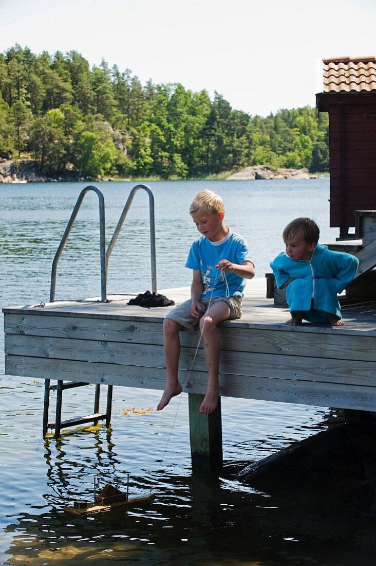 Zwei Kinder sitzen auf Holzsteg und spielen mit Holzschiffchen im See