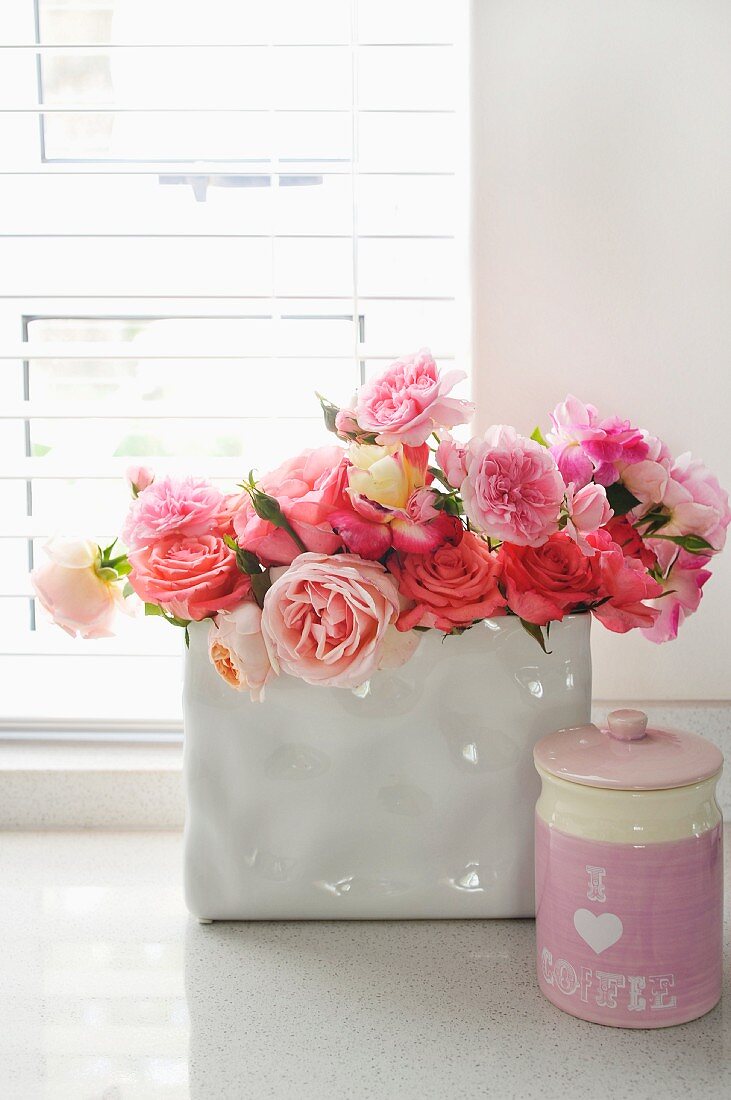 Bouquet of roses in white china vase next to pink ceramic pot with lid on windowsill