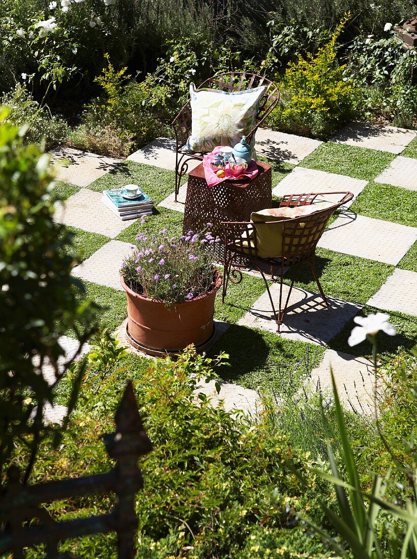 Delicate garden furniture and large terracotta planter of flowers on chequered pattern of stone slabs and squares of lawn