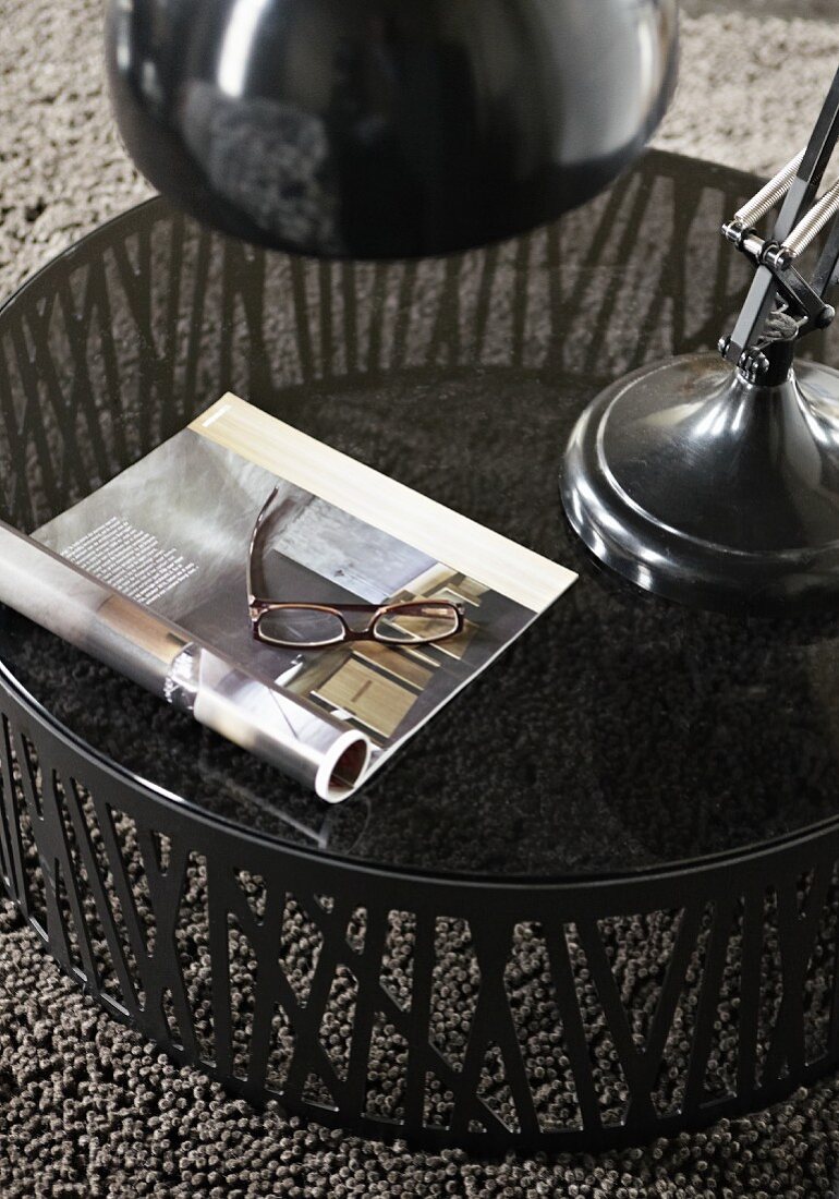 Runder Beistelltisch mit Glasplatte auf grauem Teppich; auf dem Tisch ein aufgeschlagenes Buch und eine Lesebrille
