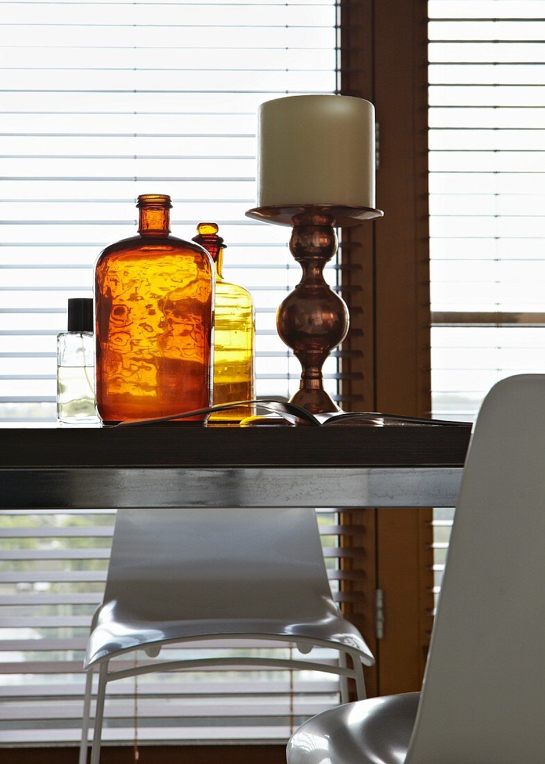 Large, antique candlestick next to brown vintage bottle on modern table in front of window