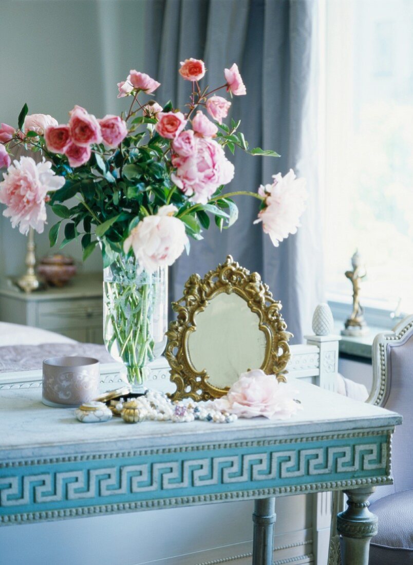 Goldgerahmter Spiegel neben duftendem Rosenstrauss auf antikem, pastellblauen Tisch mit griechischem Muster