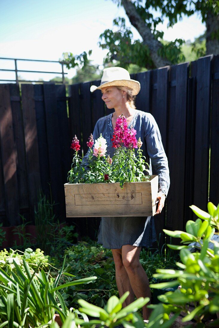 Frau im Garten trägt Holzkiste mit Blumen