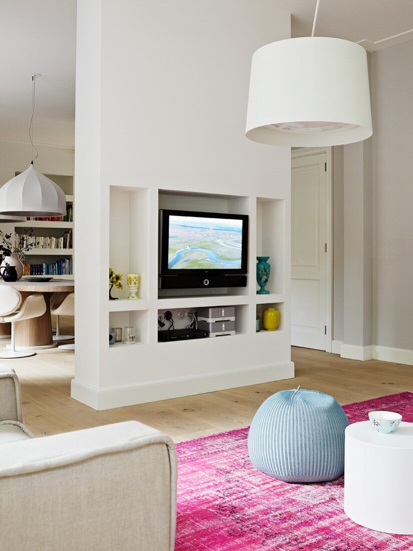 Ess- und Wohnraum mit weißem Raumteiler; in den Wandnischen finden der Fernseher und Dekorationsobjekte Platz