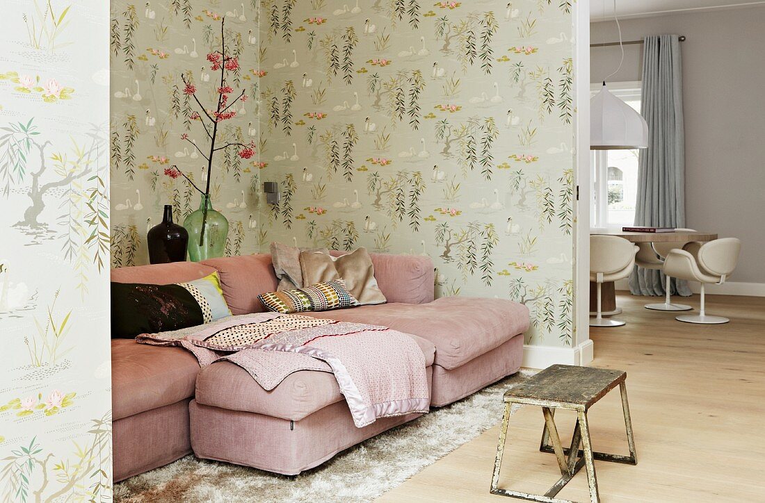 Wohnecke mit rosafarbener Couch und gemusterter Wandtapete im Retrostil; im Vordergrund ein Vintage-Tisch