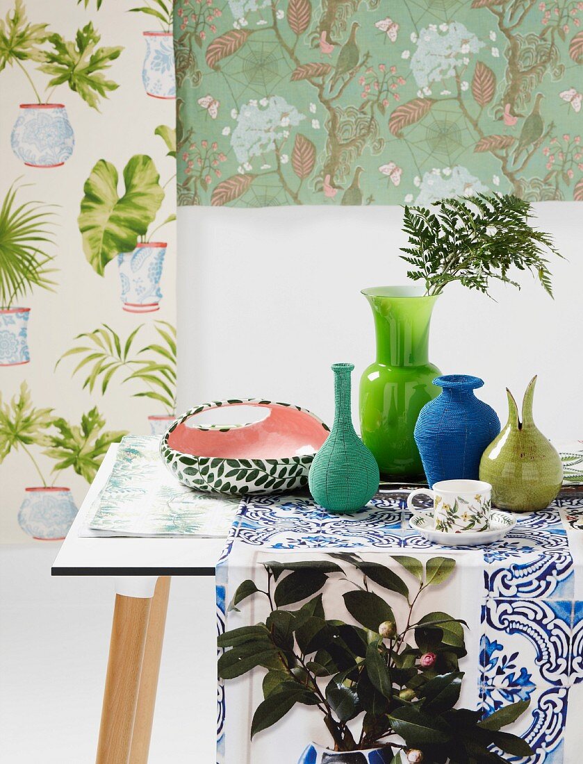 Vasensammlung in Grün- und Blautönen auf Tischläufer arrangiert vor verschiedenen Mustertapeten mit floralen Motiven
