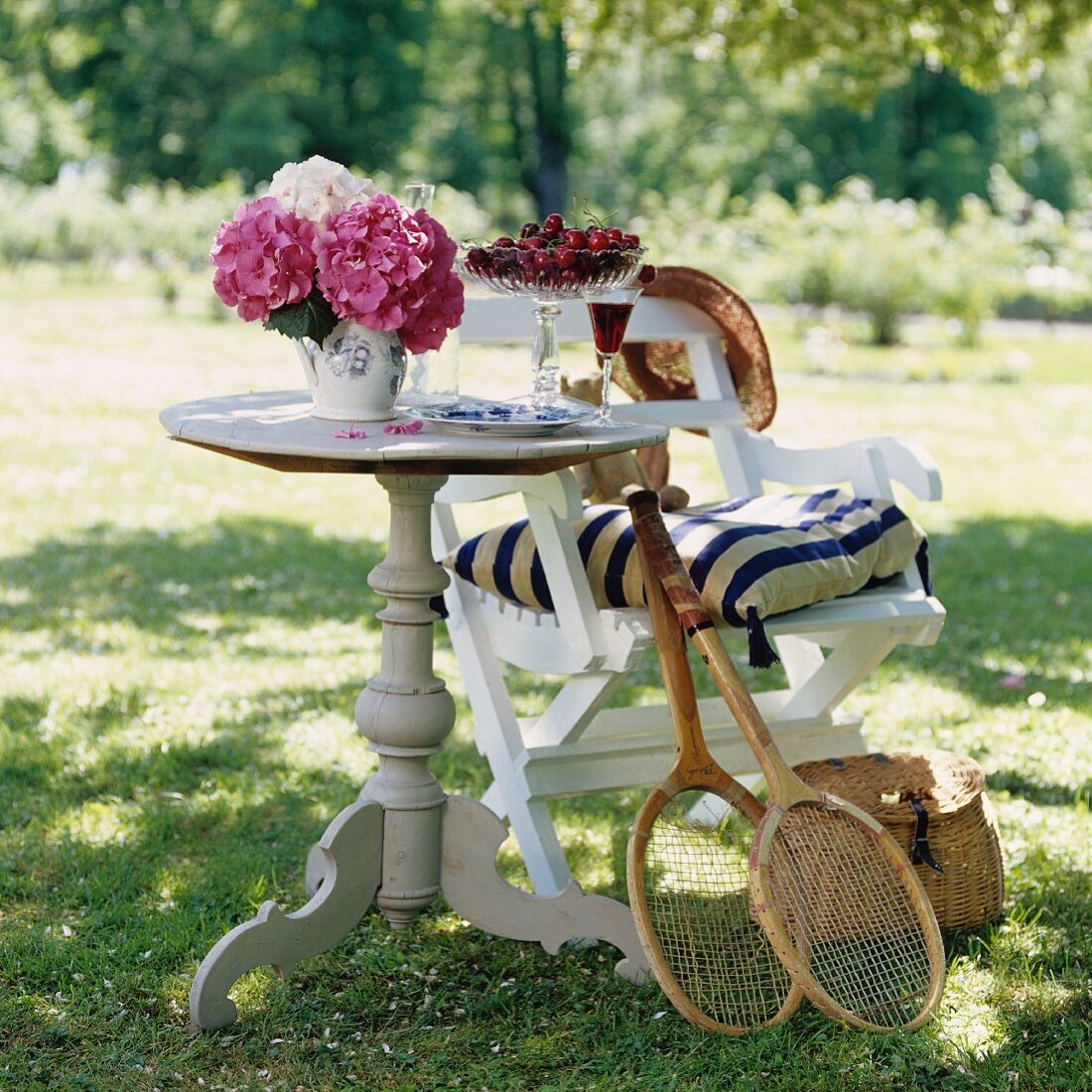 Runder Holztisch mit Kirschen und Hortensien, Tennisschläger an Stuhl gelehnt im sonnigen Garten