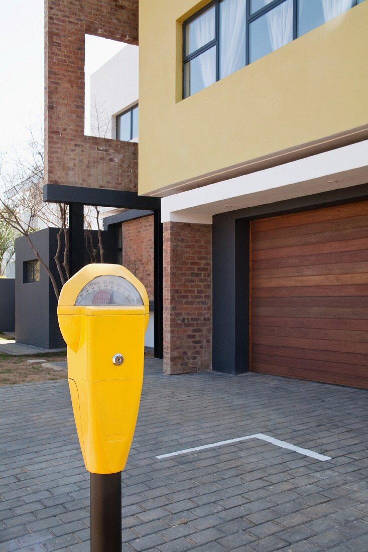 Bright yellow parking meter in front of house with garage and paved forecourt