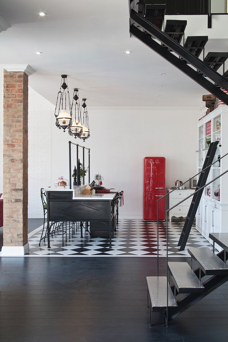 Open-plan interior with kitchen counter below group of pendant lamps and red fifties fridge on black and white floor; staircase in foreground on dark wooden floor