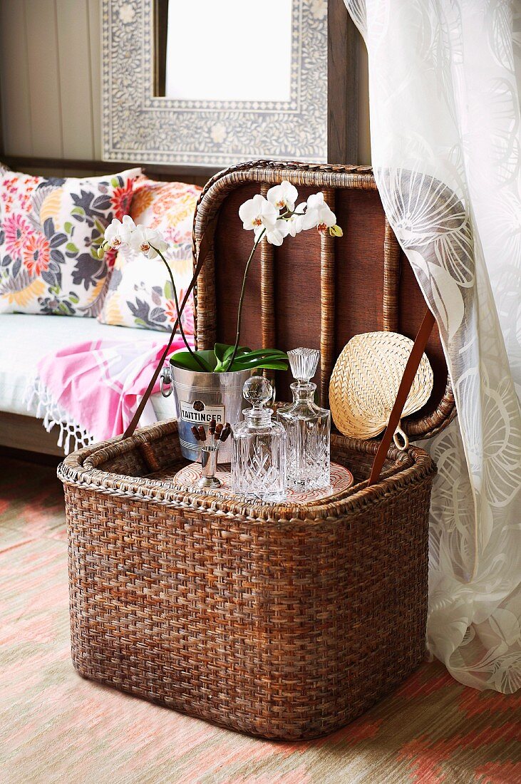 Open picnic basket with crystal carafes and orchid in metal pot; colourful scatter cushions on sofa in background