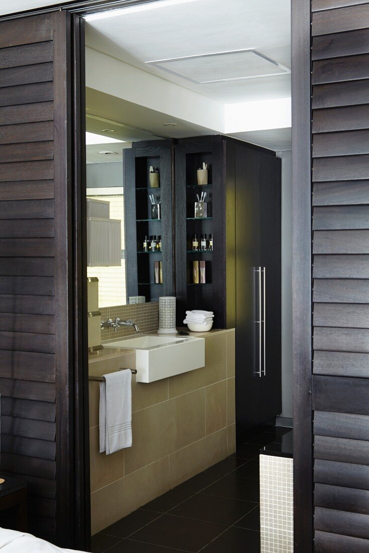 Junggesellen Bad Ensuite hinter geöffneten dunkelbraunen Schiebejalousietüren, eleganter Waschtisch mit schwarzem Einbauschrank