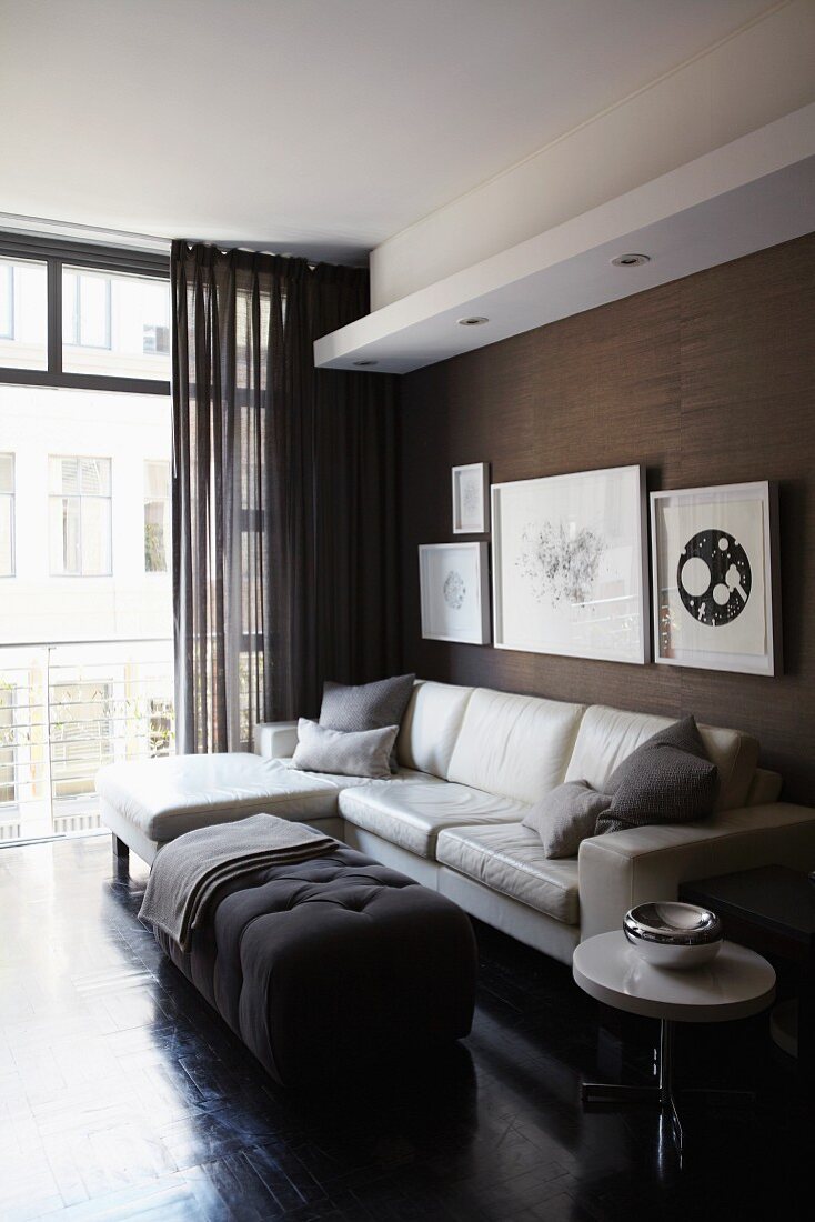Elegantes maskulines Wohnzimmer mit heller Ledercouch, gerahmten Bildern an dunkler Strukturtapete und Fensterfront