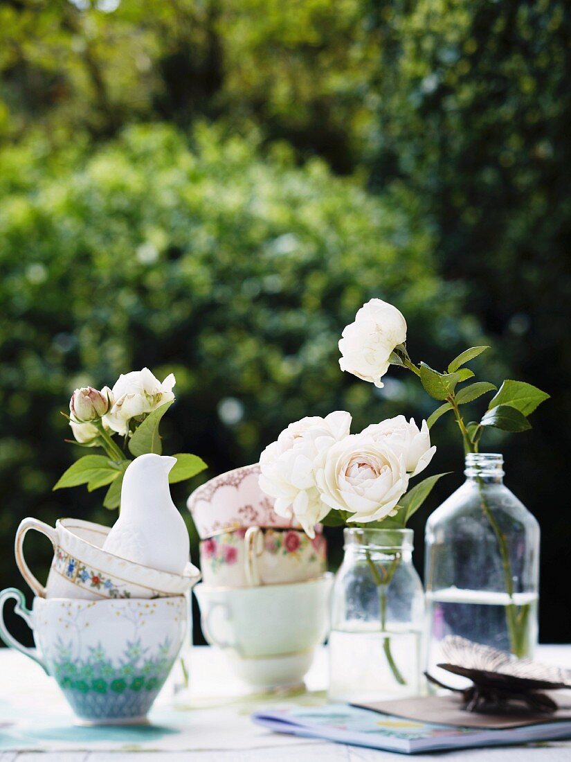 Gestapelte Vintage Tassen und weiße Rosen in Flaschen auf Tisch im Freien