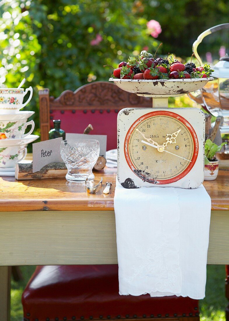 Gedeckter Tisch im sonnigen Garten; Schale mit frischen Beeren auf einer zur Uhr umgebastelten Küchenwaage