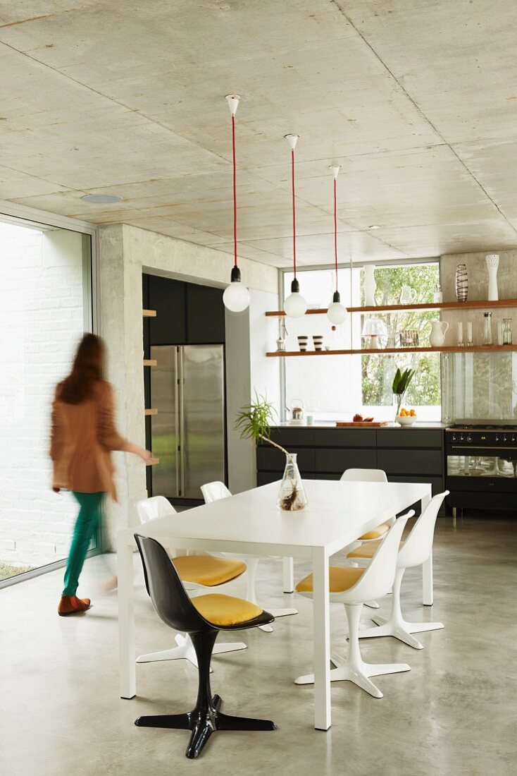 Dining table in open-plan house with exposed concrete surfaces; woman walking through room