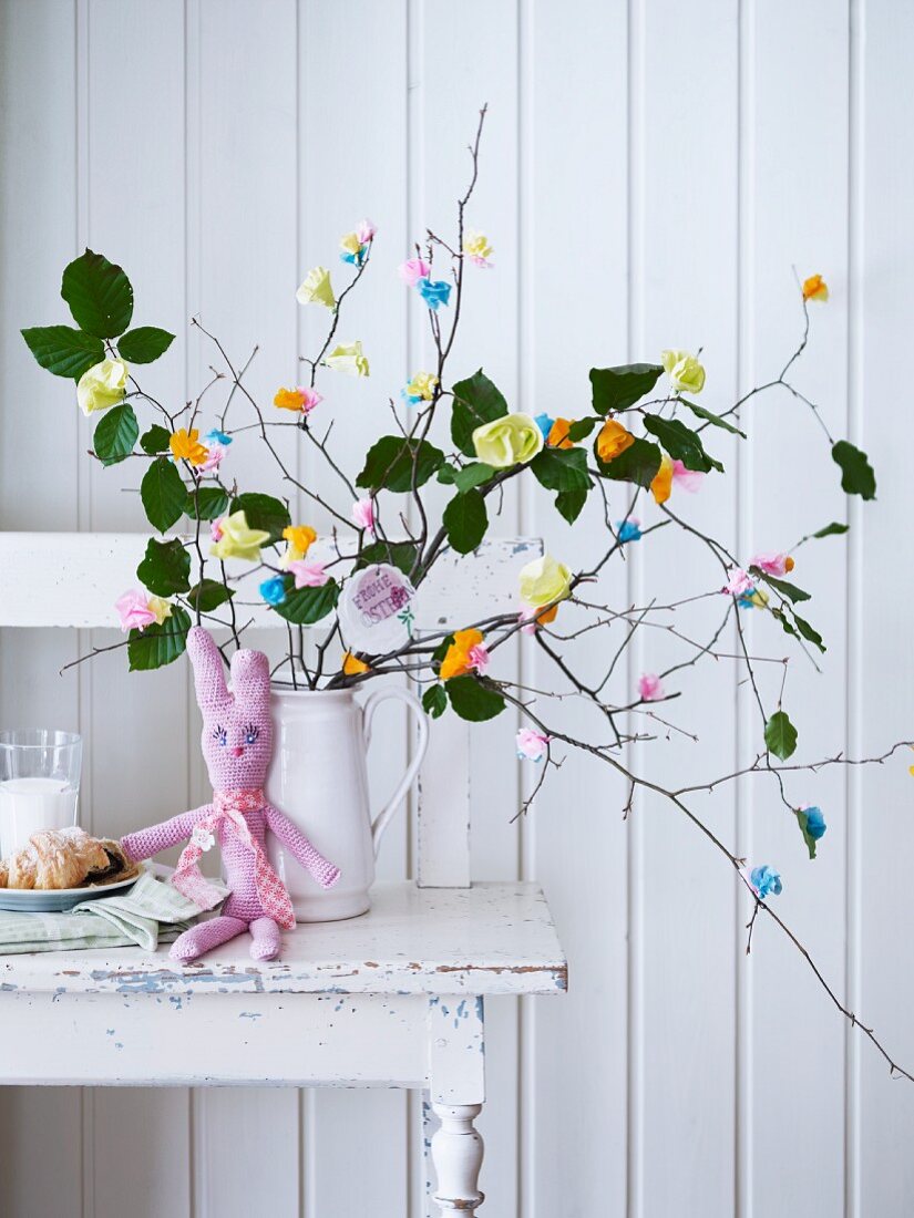 Selbstgehäkelter Osterhase vor Strauss aus Zweigen mit bunten Papierblüten dekoriert
