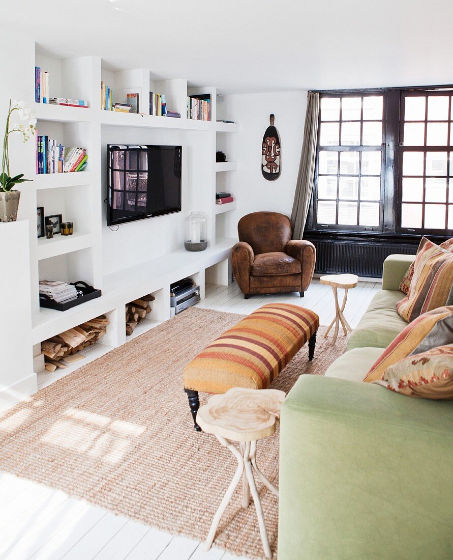 Sofa mit Kissen und gepolsterter Couchtisch gegenüber Wand mit gemauertem Regal und aufgehängtem Fernseher, im Hintergrund Ledersessel neben schwarzer Sprossentür