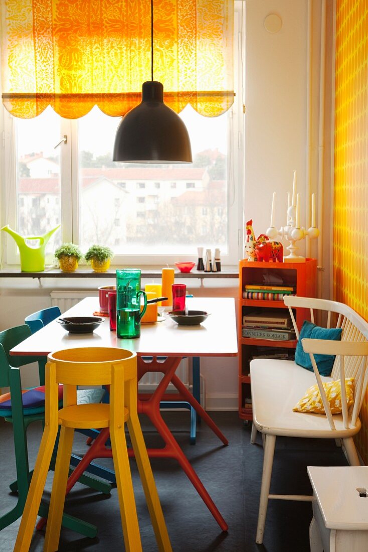 Bunt lackierte Stühle und weiße Sitzbank um Esstisch mit farbigen Gläsern, an Fenster halb geschlossenes Rollo in Gelb mit geschwungener Bordüre