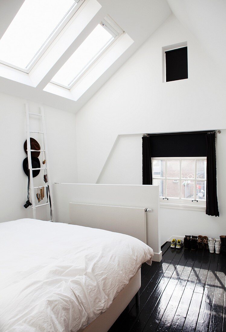 Bed with white bed linen in front of half-height balustrade wall in attic room with skylight and dark wooden floor