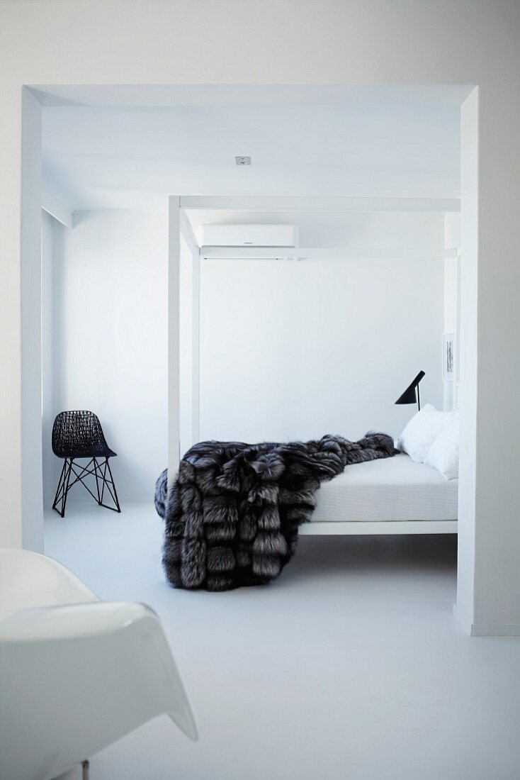 White, minimalist bedroom with fur blanket on canopied bed; Bauhaus wicker chair in corner