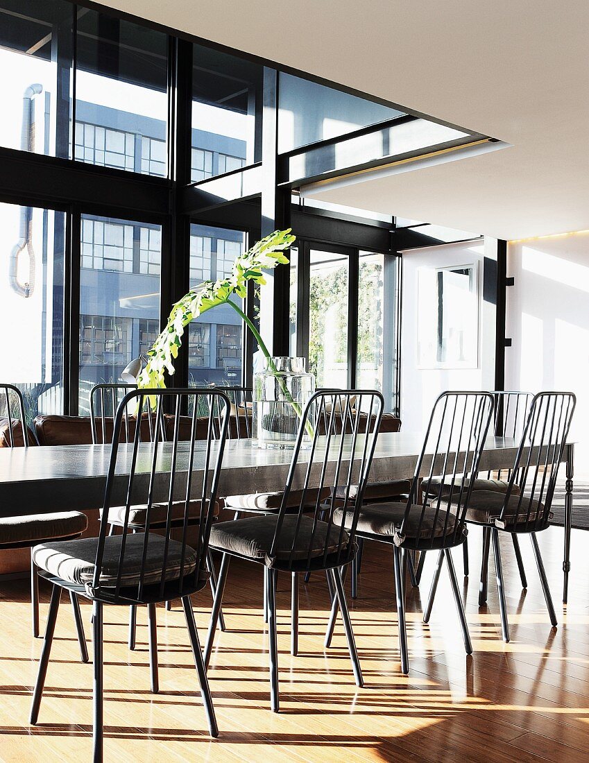Long dining table and chairs with high backrests next to steel and glass facade in penthouse apartment
