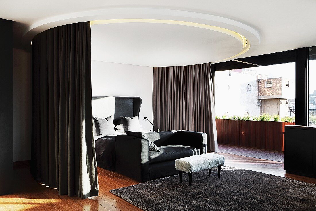 Bed and couch below circular ceiling element with integrated curtains and lights next to floor-to-ceiling window
