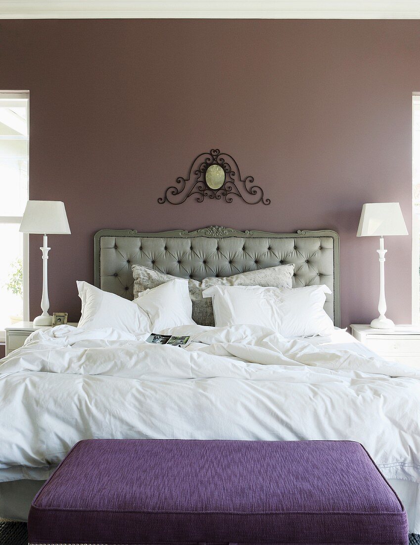 Elegantes Schlafzimmer mit gepolstertem Bettkopfteil vor aubergine getönter Pastellwand und kunsthandwerklichem Ornament