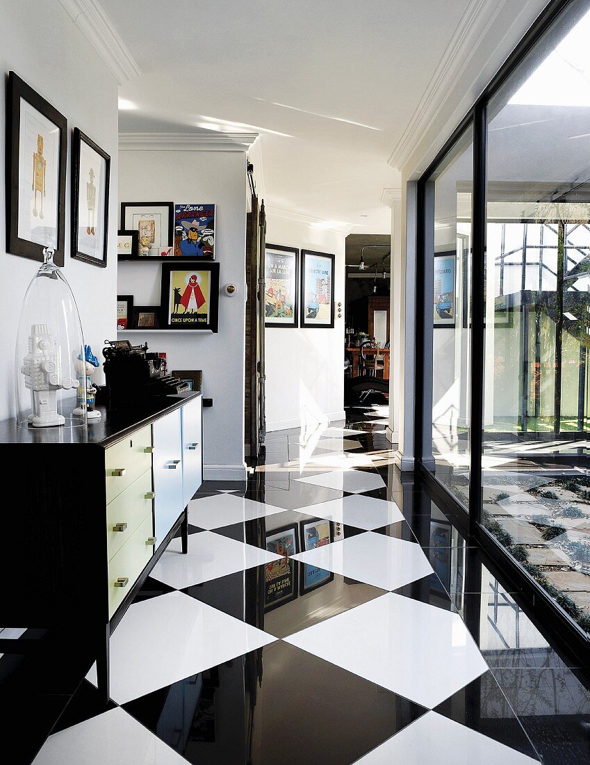 Sideboard decorated with collectors' items and gallery of pictures in hallway with elegant, chequered floor and glass wall with view of courtyard on right