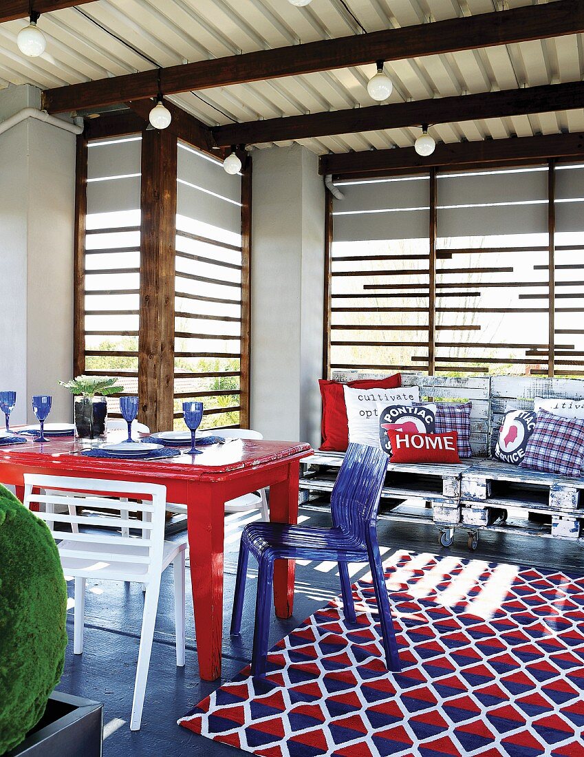 Überdachte Terrasse mit altem Tisch, Stühlesammlung und Musterteppich in Blau, Weiß und Rot, vor Sesseln aus alten Europaletten mit Kissen