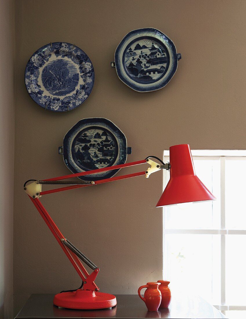 Red, metal desk lamp against wall painted pale grey with collection of traditional, blue and white plates