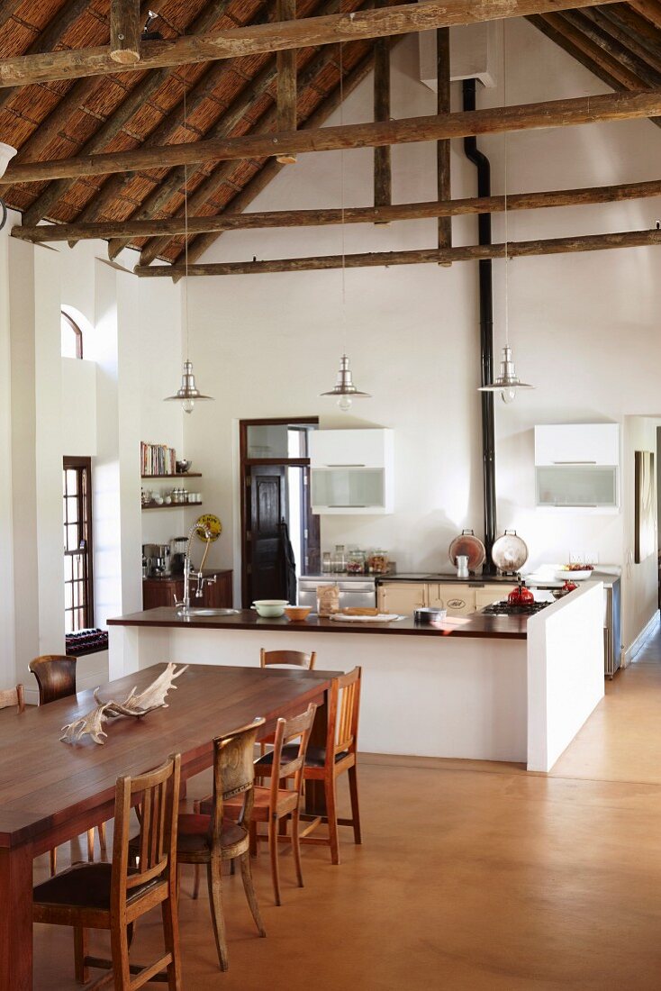 Esstisch mit verschiedenen Holzstühlen vor offener Küche mit Theke in hohem Wohnraum und Blick auf sichtbaren Dachstuhl