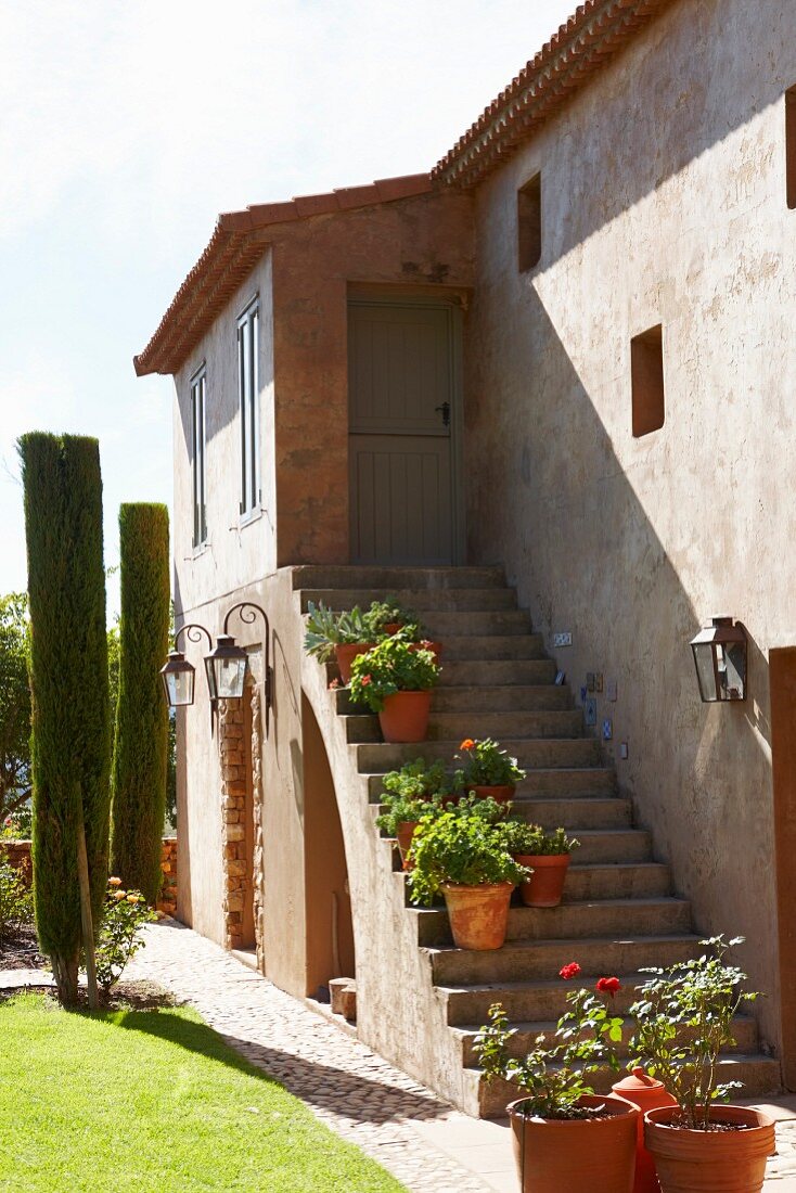 Sonnenbeschienenes Haus mit Aussentreppe und Blumentöpfen auf Stufen vor Garten mit Zypressen