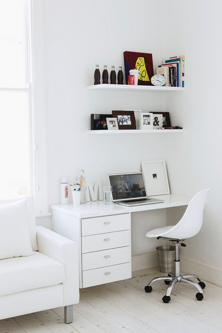 Arbeitsecke - weisser Drehstuhl vor Schreibtisch mit Schubläden und Bords an Wand neben Sessel vor Fenster