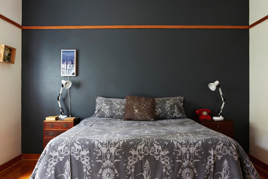 Doppelbett mit gemusterter Tagesdecke vor grau getönter Wand und umlaufender Holzleiste