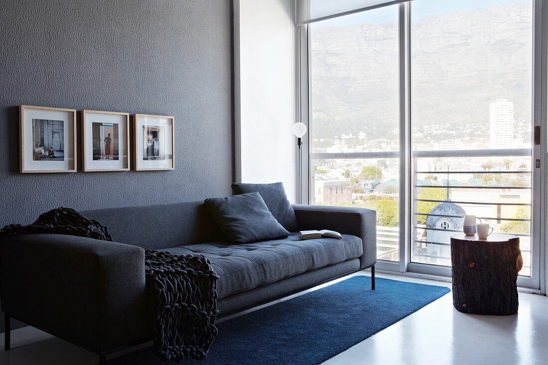 Moderne, anthrazitfarbene Couch an grauer Wand; neben der Couch eine raumhohe Glasfront mit herrlichem Blick auf das Häusermeer und die Berge