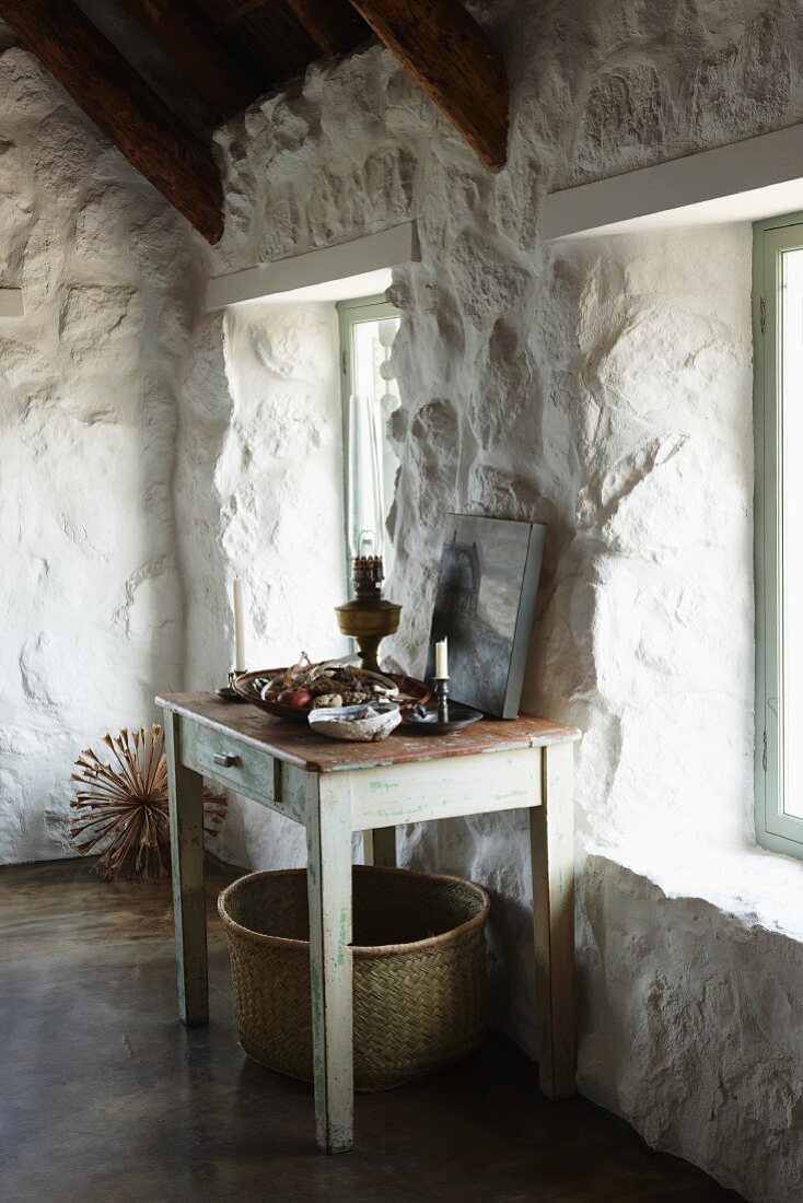 Vintage wooden table against whitewashed, rustic stone wall in pale room; large basket below table