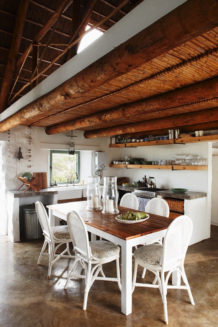 Open-plan interior with simple kitchen, wooden table and white chairs below rustic wood-beamed ceiling