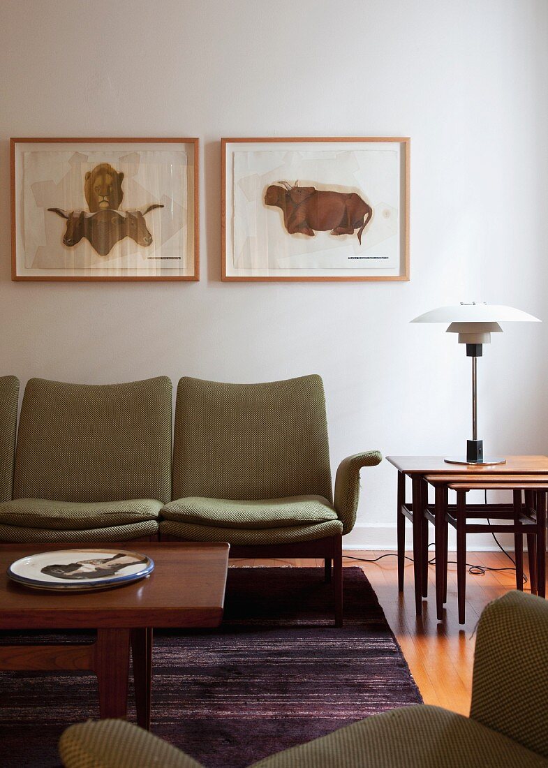 Couchtisch aus Holz vor gepolsterter Sitzbank an Wand mit gerahmten Bildern und mehrteiliger Beistelltisch mit Klassiker Tischleuchte