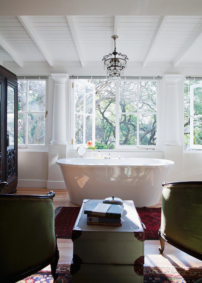 Nostalgic bathroom with modern, free-standing bathtub and two antique armchairs; stack of two antique trunks used as table