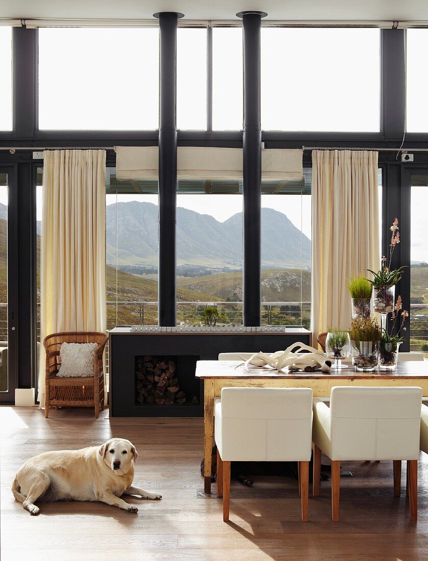 Black wood-burning stove with two stovepipes in front of magnificent mountain landscape in open-plan interior with rustic dining table and white armchairs