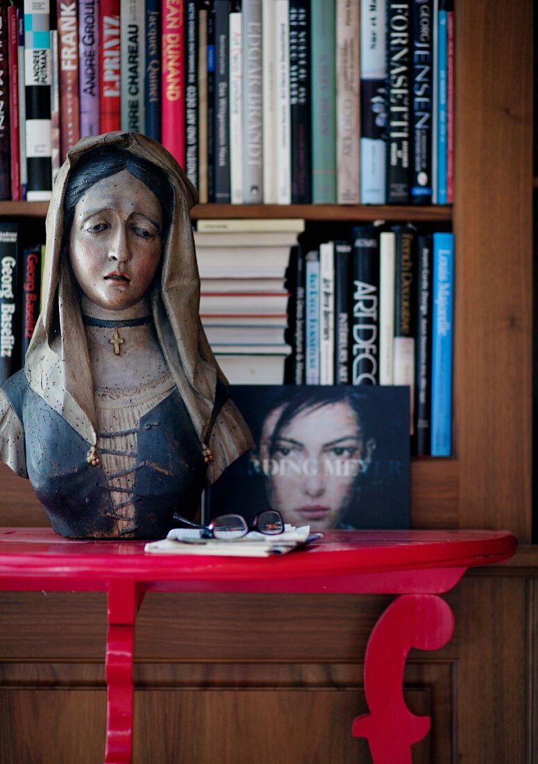 Painted, wooden bust of Madonna on red console table in front of bookcase