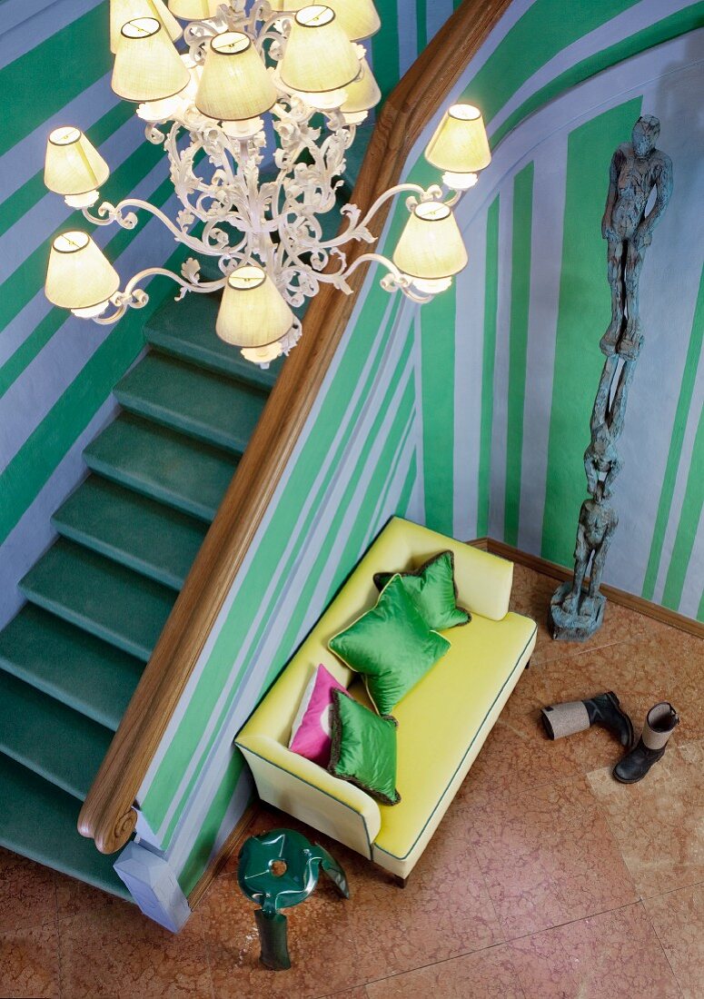Chandelier and yellow sofa in grand stairwell with blue and green striped walls; totem pole artwork (stacked human forms) on marble floor