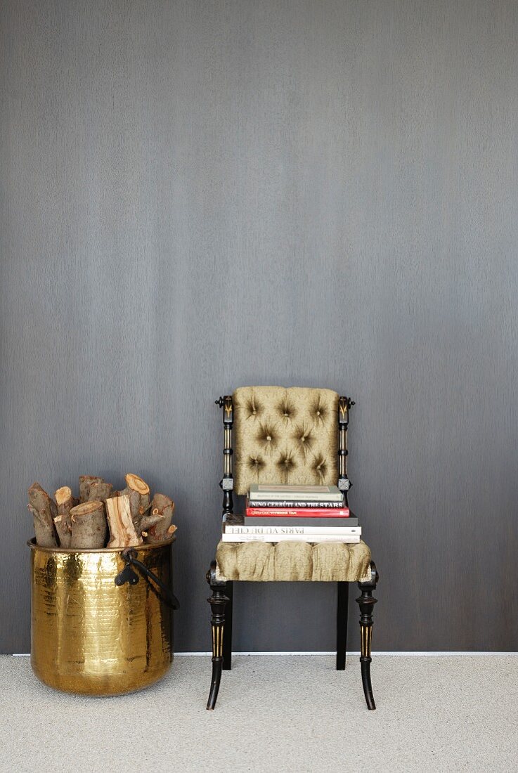 Neo-antique chair with upholstered seat and back with books stacked on seat next to brass pot of logs against grey wall