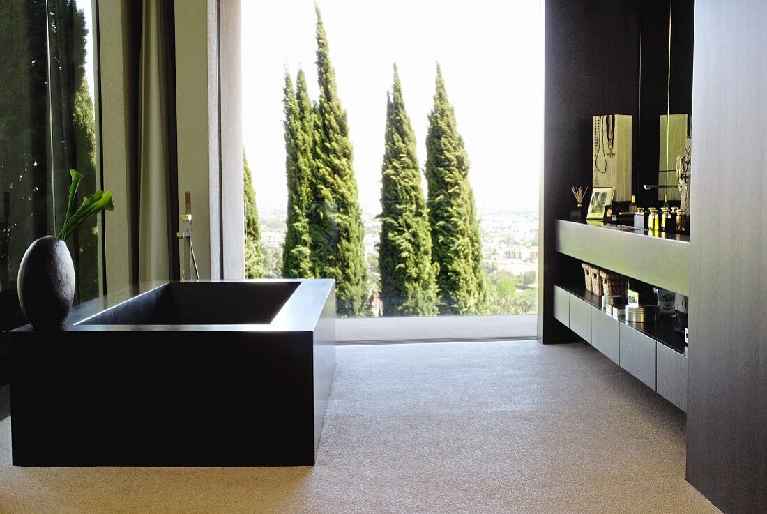 Schwarze Badewanne gegenüber eingebautem Waschtisch, im Hintergrund Fensterfront mit Blick auf Baumgruppe