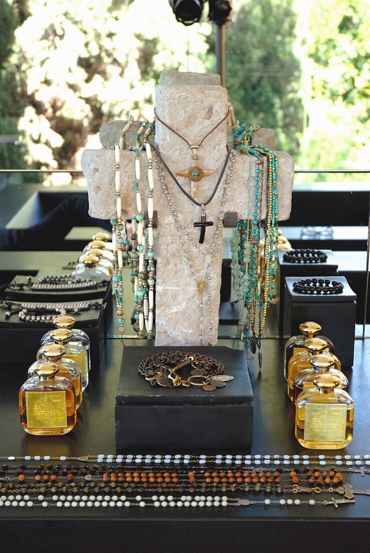 Kreuzförmiger Schmuckständer mit Halsketten behängt, davor schwarzes Kästchen, Parfumflakons und ausgebreitete Halsketten auf Ablage